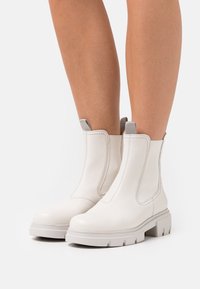 Nohy obuté do bílých kotníkových bot s robustními podešvemi a šedými poutky, stojící na obyčejném bílém pozadí.