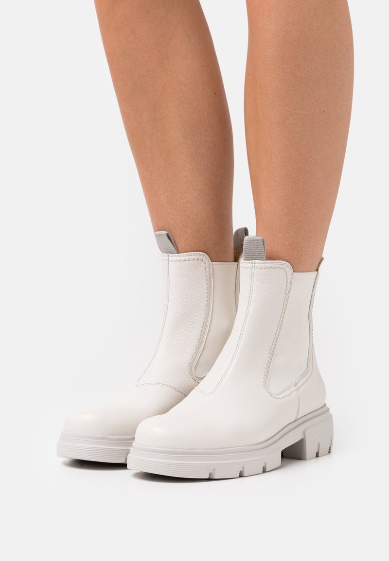 Nohy obuté do bílých kotníkových bot s robustními podešvemi a šedými poutky, stojící na obyčejném bílém pozadí.