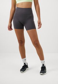 Shorts de sport taille haute gris foncé avec une texture lisse, des coutures accentuées et un design ajusté. Portés avec des baskets noires et des chaussettes blanches.