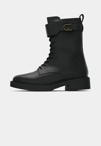 Bottines noires à lacets en cuir lisse avec une semelle plate et épaisse, et un accent de sangle avec une boucle dorée.