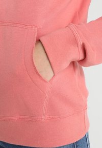 Korálový svetr s klokaní kapsou a ribovanými manžetami. Měkká, texturovaná tkanina s viditelným švem podél švů.