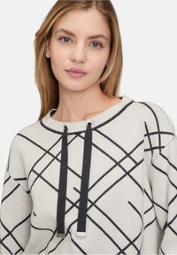 Helles beige Sweatshirt mit schwarzen abstrakten Linien, weitem Halsausschnitt und dunklen Kordeln mit silbernen Metallspitzen. Weiche Textur.
