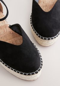 Schwarze Wildleder Keil-Espadrilles mit Knöchelriemen und gewebten Jutesohlen auf hellem Hintergrund.