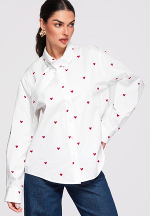 Femme portant une chemise blanche à boutons avec un motif de petits cœurs rouges, associée à un jean bleu foncé, debout avec une main sur la hanche.