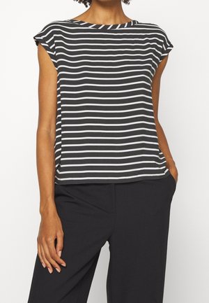 Vrouw draagt een zwart-wit horizontaal gestreept mouwloos topje en een zwarte broek, met één hand in haar zak.