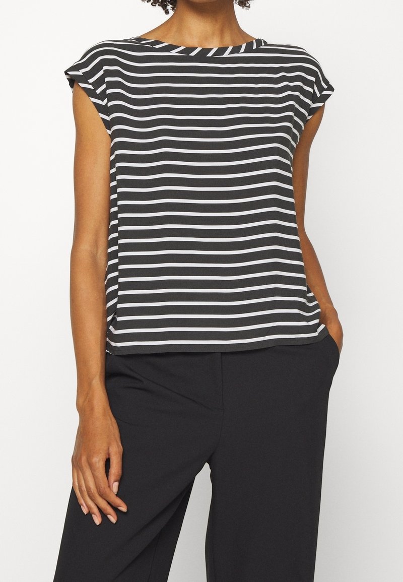 Femme portant un haut sans manches à rayures horizontales noires et blanches et un pantalon noir, une main dans la poche.