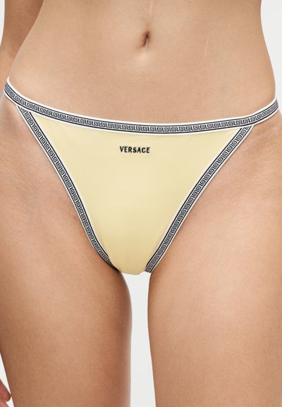 Bas de bikini jaune avec bordure géométrique noire et blanche et logo Versace centré à l'avant, porté sur peau nue.
