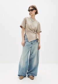 Kvinde iført oversized lyseblå jeans, beige top med rynket side, brune flade sko med nitter, solbriller og sølv kæde halskæde mod hvid baggrund.