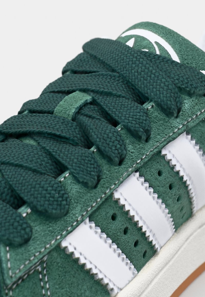 Grüne Wildledersneaker mit weißen Lederstreifen, strukturierten grünen Schnürsenkeln, perforierten Abschnitten und einer stabilen weißen Gummisohle.