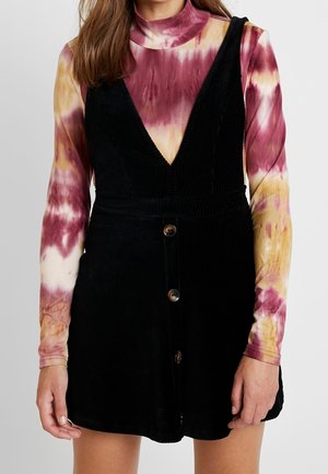 Nainen, joka käyttää mustaa puuvillaisen mustekankaasta valmistettua napitettavaa essumekkoa pitkähihaisen, puna-, keltavärisen ja kermaan vivahtavan tie-dye-kuvioisen paidan päällä.