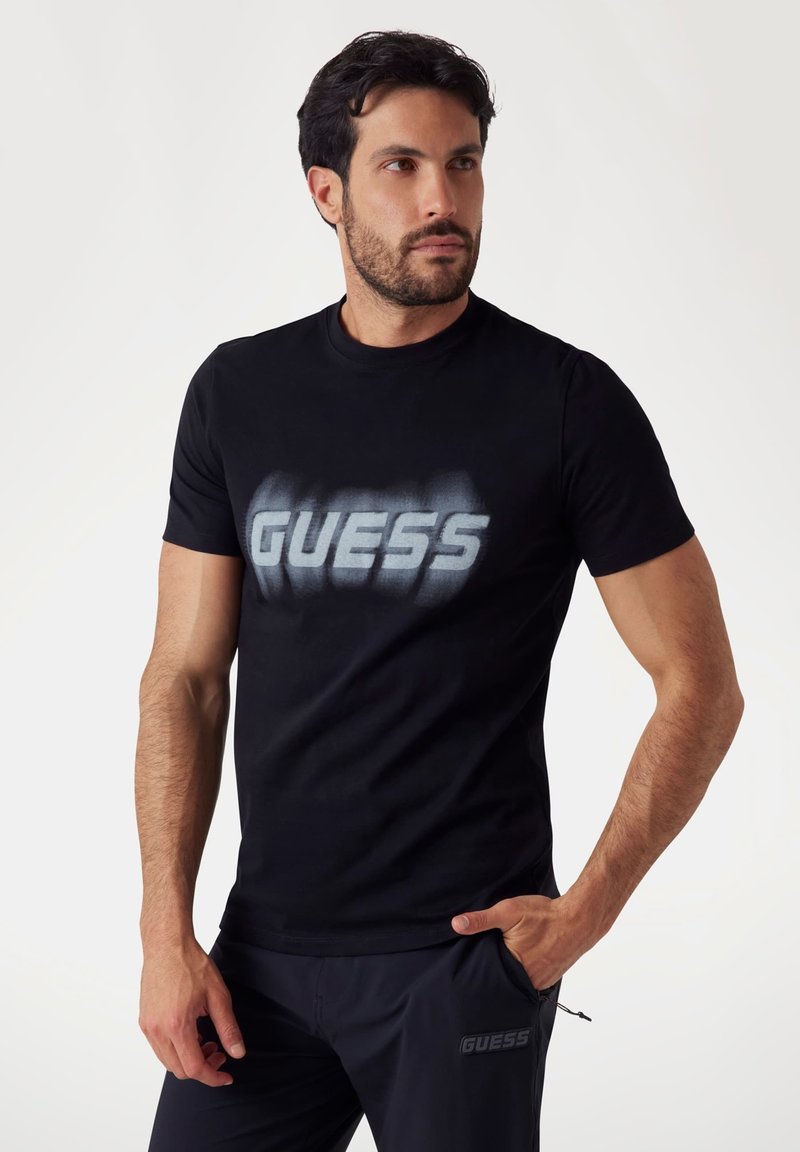 Camiseta de algodón negra con un gran logo en gris desgastado de "GUESS" en el frente. Manga corta y diseño clásico de cuello redondo.