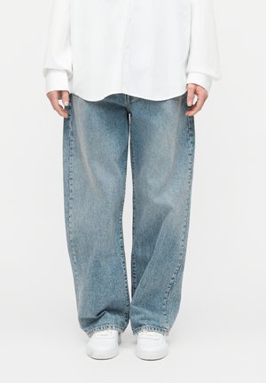 Osoba v svetlomodrých džínsach s širokými nohavicami, bielej košeli na gombíky a bielych teniskách, stojaca pred jednofarebným pozadím.