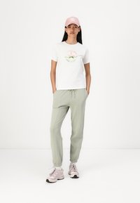 Witte T-shirt met kleurrijk "Converse All Star" logo, lichtgroene joggers en roze pet. Model draagt lichtroze sneakers met grijze accenten.