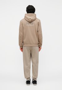 Beige hoodie met een trekkoord, voorzien van een logo op de achterkant, gecombineerd met bijpassende sweatpants. Materiaal heeft een zachte, gestructureerde afwerking.