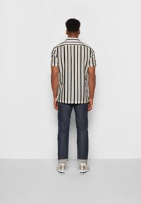 Korte mouwen shirt met verticale zwarte strepen op een lichte achtergrond, gecombineerd met donkere denim broek en sneakers, gezien van achteren.