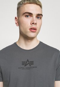 Pilkos spalvos medvilnės marškinėliai su apvaliu iškirpimu ir trumpomis rankovėmis, ant krūtinės atspausdintu juodu logotipu. Lygi tekstūra.