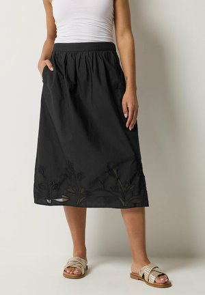 Kobieta ubrana w czarną spódnicę midi z kwiatową haftem przy krawędzi, biały top na ramiączkach i beżowe plecione sandały, stojąca na jednolitym tle.