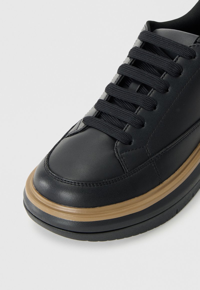 Zapatilla negra de cuero con punta redonda, media suela marrón en contraste y cordones negros. Presenta una textura suave y un diseño sencillo.