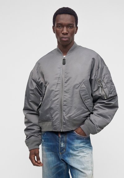 Jeune homme portant une veste bomber grise avec une fermeture éclair avant et un jean bleu délavé, debout devant un fond clair uni.