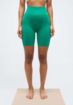 Femme debout pieds nus sur un tapis beige, portant un short de cycliste côtelé vert taille haute, visible de la taille aux pieds.