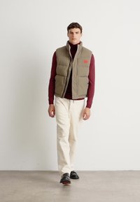 Man walking in beige padded vest, burgundy turtleneck, white pants, and black shoes against plain white wall and concrete floor.