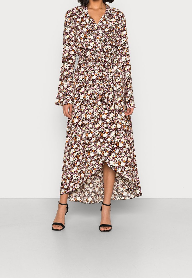 Vestido maxi floral con mangas largas, escote en V y cintura anudada, en tonos de marrón, blanco y naranja con una silueta fluida. Tacones negros.