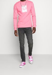 Pink long-sleeve shirt with graphic print on the front, paired with dark jeans and patterned pink sneakers.