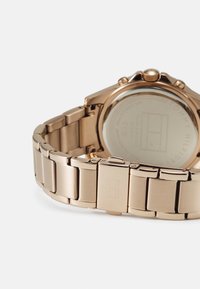 Montre en acier inoxydable doré rose avec un bracelet poli et un logo gravé sur le fermoir. Le fond du boîtier présente des marquages de la marque.