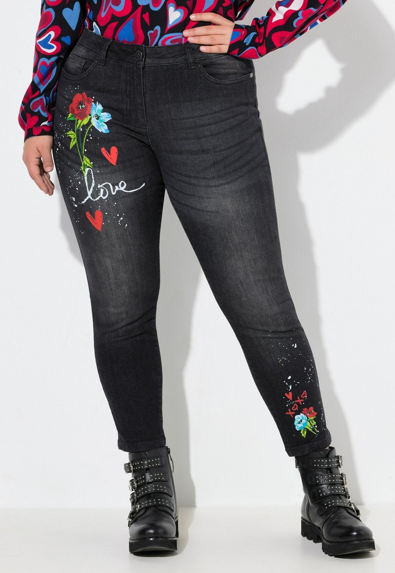 Jeans skinny negros con bordados florales en rojo y azul, que presentan corazones y la palabra "amor". Incluye detalles de salpicaduras blancas y costuras visibles.
