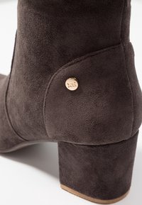 Botín de ante marrón hasta el tobillo con punta redonda, superficie texturizada y un acento de logo en tono dorado en el costado. Diseño de tacón bloque.