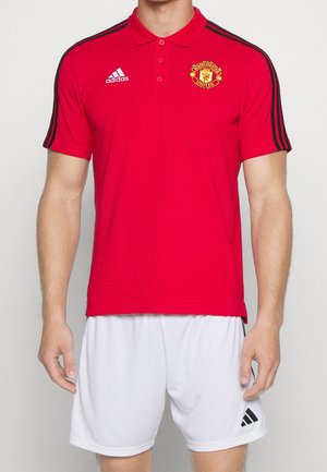Homme portant un polo rouge de Manchester United et un short blanc Adidas, debout avec les bras détendus le long du corps.