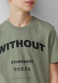 Olivgrünes Baumwoll-T-Shirt mit auffälligem schwarzen Text. Auf der Front steht das Wort "OHNE" sowie zusätzlicher Text in Englisch und Japanisch. Kurzarm.