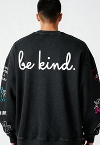 Črna oversize pulover z napisom "bodite prijazni." v belem pisavi na hrbtni strani. Okrašen s pisanimi grafikami na rokavih. Teksturna tkanina.