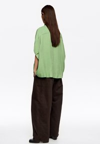 Camisa verde, holgada y de manga corta con una textura sutil, combinada con pantalones amplios de color marrón oscuro. Diseño simple y sin adornos.