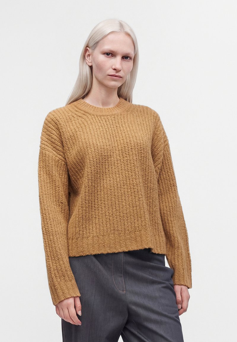 Jeune femme aux longs cheveux blonds portant un pull marron texturé et un pantalon gris foncé sur un fond blanc uni.