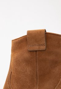 Bottine en daim marron avec un onglet cousu en haut, texture lisse et détails de coutures courbes sur les côtés.