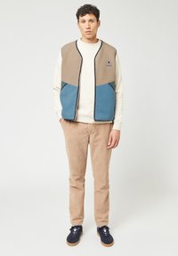 Fleece vest met kleurblokontwerp in beige, teal en donkergroen. Draag het over een crèmekleurige trui, gecombineerd met beige corduroy broek en marineblauwe schoenen.