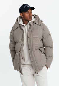 Bruin en wit geruit pufferjack met een capuchon, voorzien van een centrale rits en drukknoopjes. Draagt over een lichtgrijze hoodie.