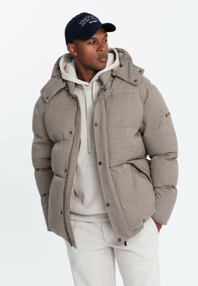 Bruin en wit geruit pufferjack met een capuchon, voorzien van een centrale rits en drukknoopjes. Draagt over een lichtgrijze hoodie.