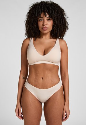 Vrouw met krullend haar die een beige beha en ondergoed draagt, staand tegen een effen lichtgrijze achtergrond, kijkend naar voren.
