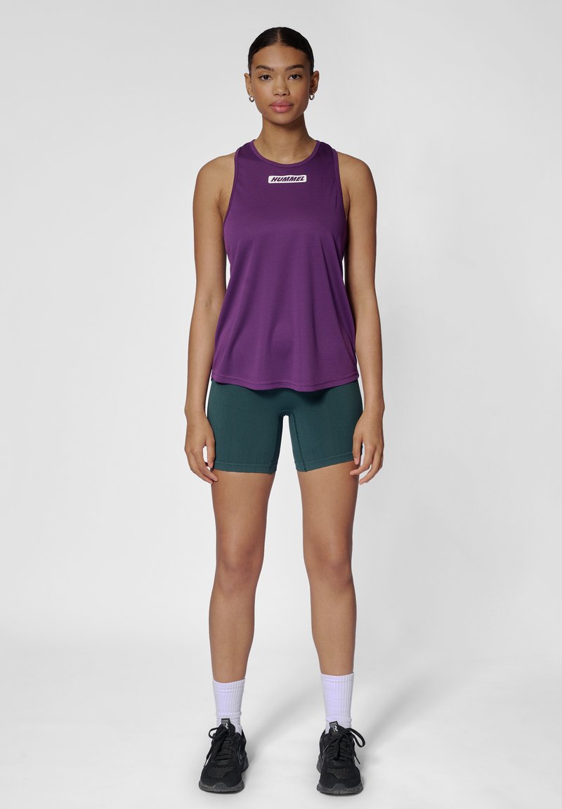Kobieta stoi twarzą do przodu, ubrana w fioletowy sportowy top bez rękawów, ciemnozielone krótkie spodenki, białe skarpetki i czarne buty do biegania.