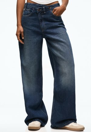 Persoon die donkerblauwe brede jeans draagt met twee knopen in de taille en witte sneakers, staand met één hand in de zak.