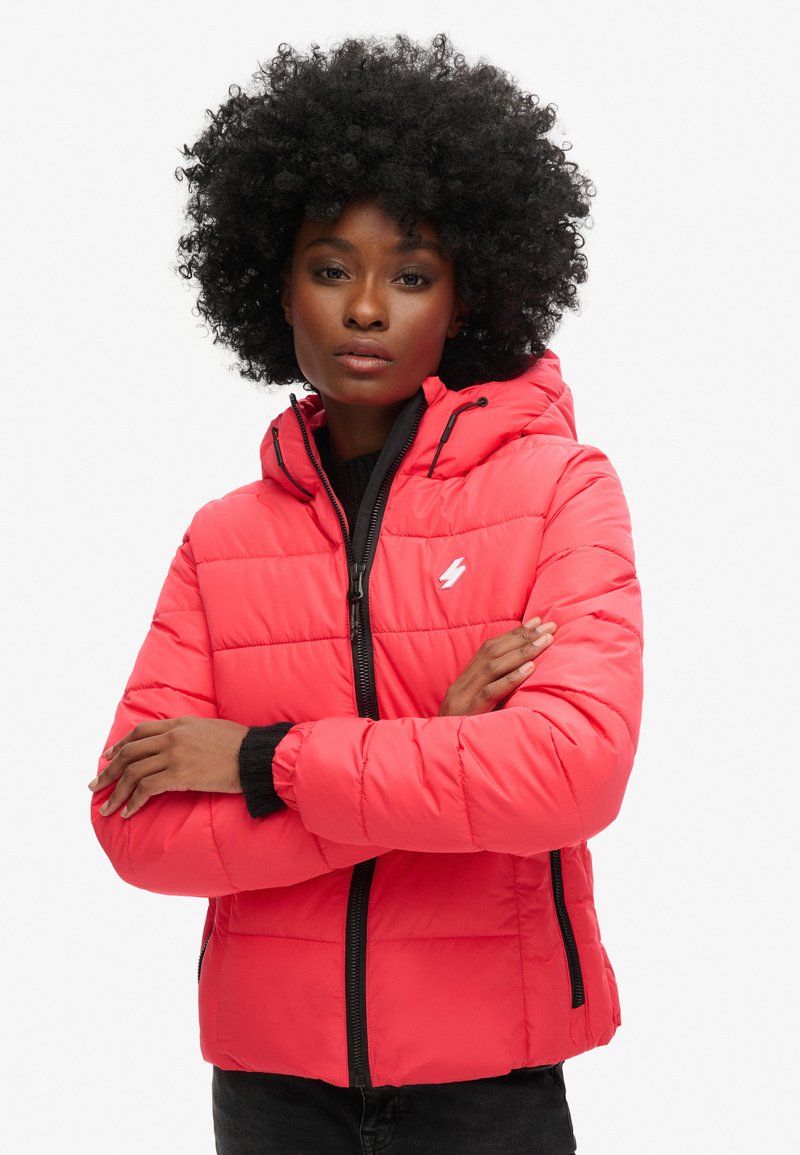 Czerwona pikowana kurtka z zamkiem z przodu, regulowanym kapturem i czarnymi akcentami. Zawiera pikowaną teksturę oraz logo po lewej stronie klatki piersiowej.