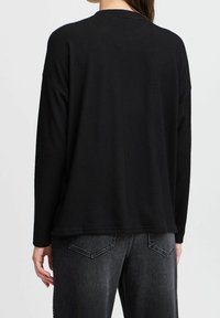 Schwarzes Langarmoberteil aus Waffelstrick mit runder Ausschnittkante, fallenden Schultern und lockerer Passform, kombiniert mit dunkelgrauen Jeans mit Taschendetails.