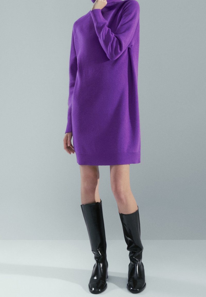 Personne portant une longue robe pull col roulé violette et des bottes noires brillantes montantes jusqu'aux genoux, debout devant un fond gris neutre.
