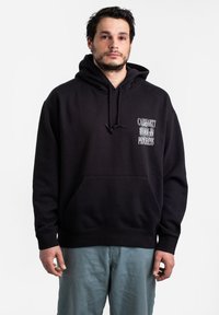 Hoodie di cotone nero con tasca frontale, cappuccio con cordino e testo stampato bianco sul lato sinistro del petto. Indossato con pantaloni azzurri chiari.