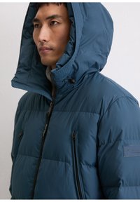 Doudoune bleue avec une grande capuche, dotée d'une fermeture éclair à l'avant et de deux poches poitrine zippées. Texture lisse et matelassée avec détail de logo.