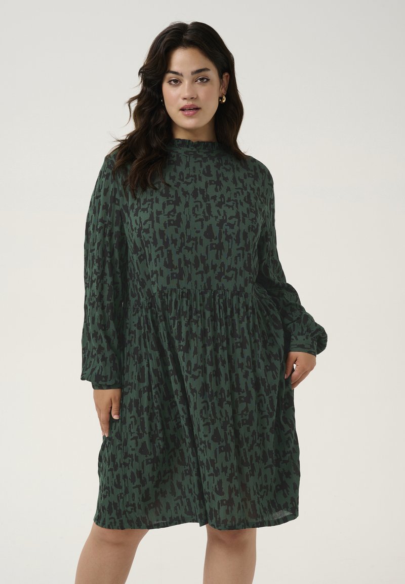Robe verte avec un motif abstrait noir, manches longues, col haut et taille froncée, fabriquée en tissu léger.
