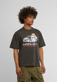 Pelēka īsām piedurknēm t-kreklā ir attēlots karikatūras lācītis un sacīkšu automobilis ar tekstu "MARKET Ultimate Performance" baltā un sarkanā krāsā.