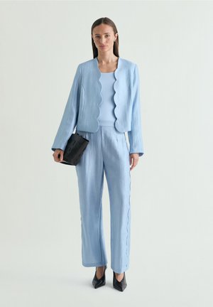 Vrouw draagt een lichtblauw pak met geschulpte rand en zwarte hakken, houdt een zwarte gestructureerde clutch vast, staand tegen een effen lichte achtergrond.